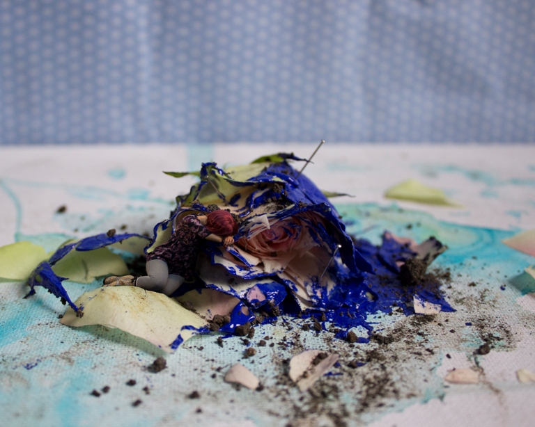 A minute woman curls up next to a rose that has been splashed with blue paint